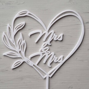 Op de afbeelding: Een witte taarttopper met de woorden "Mrs & Mrs" in een hartvormige lijst. Een decoratief bladontwerp siert de linkerkant. De topper is gemaakt van wit materiaal en heeft een kleine steel aan de onderkant.