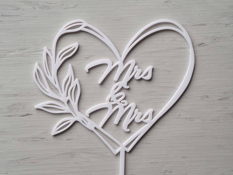 Op de afbeelding: Een witte taarttopper met de woorden "Mrs & Mrs" in een hartvormige lijst. Een decoratief bladontwerp siert de linkerkant. De topper is gemaakt van wit materiaal en heeft een kleine steel aan de onderkant.