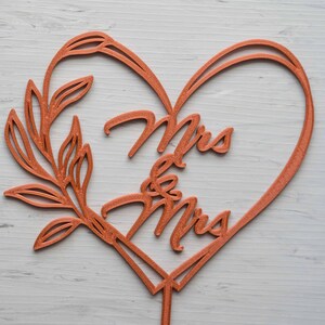 Op de afbeelding: Een koperkleurige taarttopper voor bruiloften met de woorden "Mrs & Mrs" in een hartvormige lijst. Aan de linkerkant van het hart bevindt zich een decoratief bladontwerp. De topper is bedoeld om in een taart te worden gestoken.