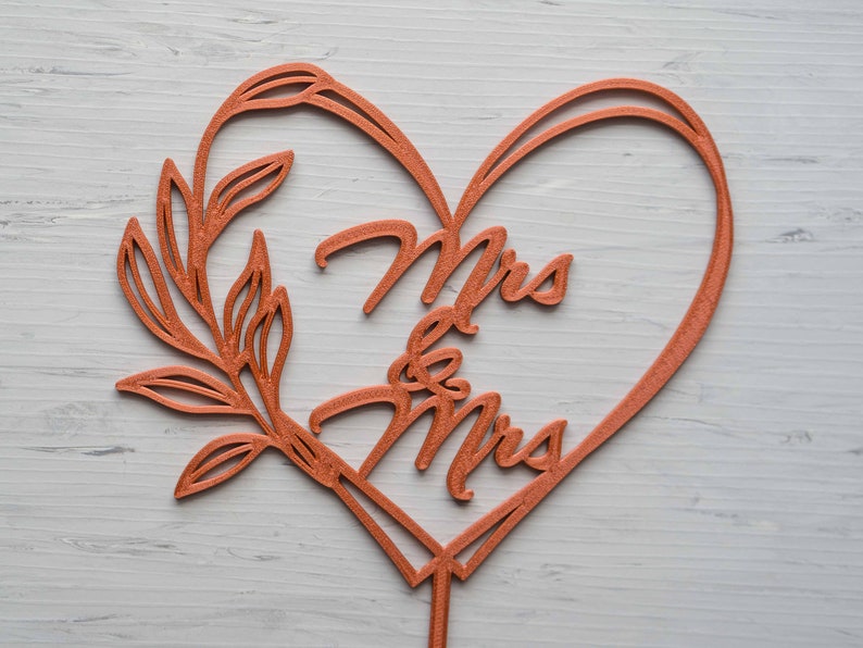 Op de afbeelding: Een koperkleurige taarttopper voor bruiloften met de woorden "Mrs & Mrs" in een hartvormige lijst. Aan de linkerkant van het hart bevindt zich een decoratief bladontwerp. De topper is bedoeld om in een taart te worden gestoken.