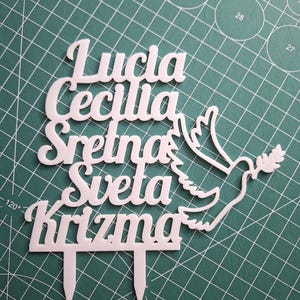 May include: A white cake topper featuring the text "Lucia Cecilia Sretna Sveta Krizma" in a cursive font, accompanied by a dove silhouette holding an olive branch. The topper includes two pointed stakes at the bottom for insertion into a cake.