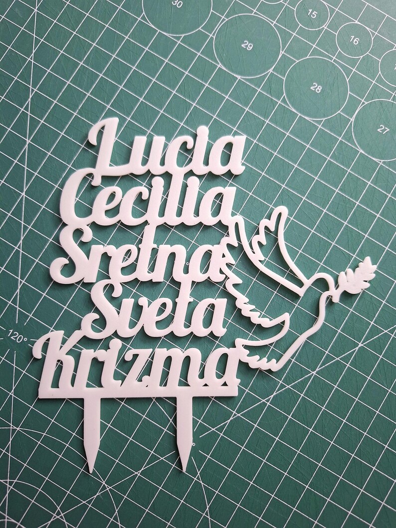 May include: A white cake topper featuring the text "Lucia Cecilia Sretna Sveta Krizma" in a cursive font, accompanied by a dove silhouette holding an olive branch. The topper includes two pointed stakes at the bottom for insertion into a cake.