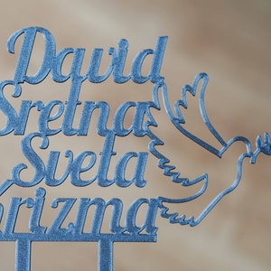 May include: A blue glittery cake topper featuring the name David and the text Sretna Sveta Krizma, accompanied by a dove silhouette holding an olive branch. The design is cut from a single piece of material with two prongs at the base.