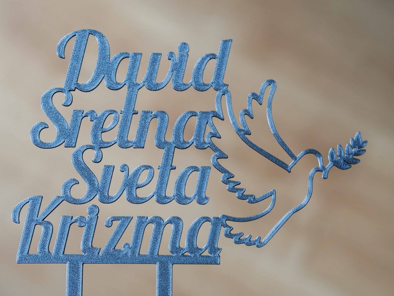May include: A blue glittery cake topper featuring the name David and the text Sretna Sveta Krizma, accompanied by a dove silhouette holding an olive branch. The design is cut from a single piece of material with two prongs at the base.
