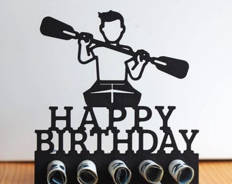 May include: A black silhouette cake topper featuring a person kayaking with a paddle above the text "HAPPY BIRTHDAY". The base includes five circular openings designed to hold rolled paper currency, creating a functional decorative display.