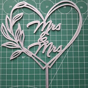 Op de afbeelding: Een zilverkleurige taarttopper met de woorden "Mrs & Mrs" in een hartvorm. Het ontwerp bevat een bloemmotief en een puntige basis. De achtergrond is een groene snijmat met metingen in centimeters.