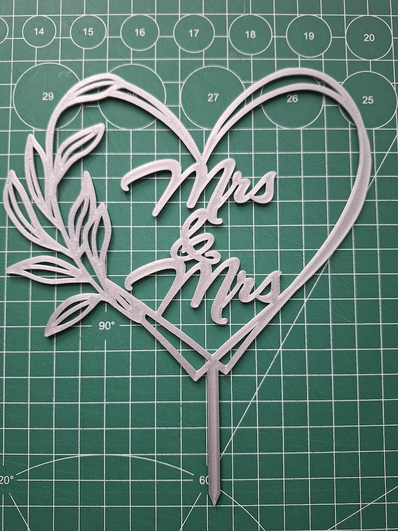 Op de afbeelding: Een zilverkleurige taarttopper met de woorden "Mrs & Mrs" in een hartvorm. Het ontwerp bevat een bloemmotief en een puntige basis. De achtergrond is een groene snijmat met metingen in centimeters.