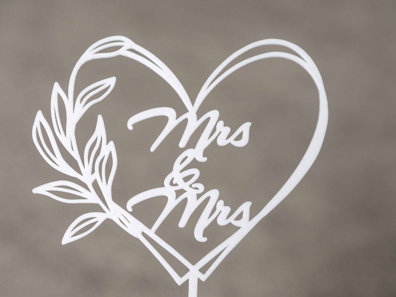 Op de afbeelding: Witte lasergesneden taarttopper met de woorden "Mrs & Mrs" in een hartvormig ontwerp. Een decoratieve bladertak bevindt zich aan de linkerkant van het hart. De topper is gemonteerd op een dunne, witte stok.