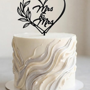 May include: A round white cake with textured, wavy silver and white frosting swirls, topped with a black cake topper shaped like a heart with a leafy branch on the left side and the text Mrs & Mrs written in the center of the heart shape.
