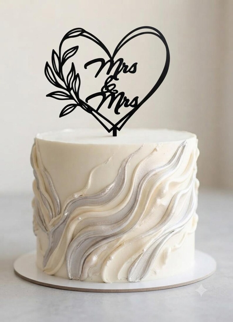 May include: A round white cake with textured, wavy silver and white frosting swirls, topped with a black cake topper shaped like a heart with a leafy branch on the left side and the text Mrs & Mrs written in the center of the heart shape.