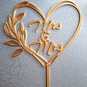 Op de afbeelding: Goudkleurige taarttopper met de woorden "Mrs & Mrs" in een hartvormige lijst. Een bladertakontwerp bevindt zich aan de linkerkant van het hart. De topper heeft een puntige stok om in een taart te steken.