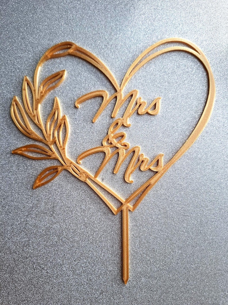 Op de afbeelding: Goudkleurige taarttopper met de woorden "Mrs & Mrs" in een hartvormige lijst. Een bladertakontwerp bevindt zich aan de linkerkant van het hart. De topper heeft een puntige stok om in een taart te steken.