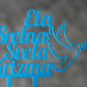 May include: A bright blue cake topper featuring the text "Ela Sretna Sveta Krizma" in a cursive font, accompanied by a stylized outline of a dove holding an olive branch. The design is cut from a single piece of material with two bottom prongs.
