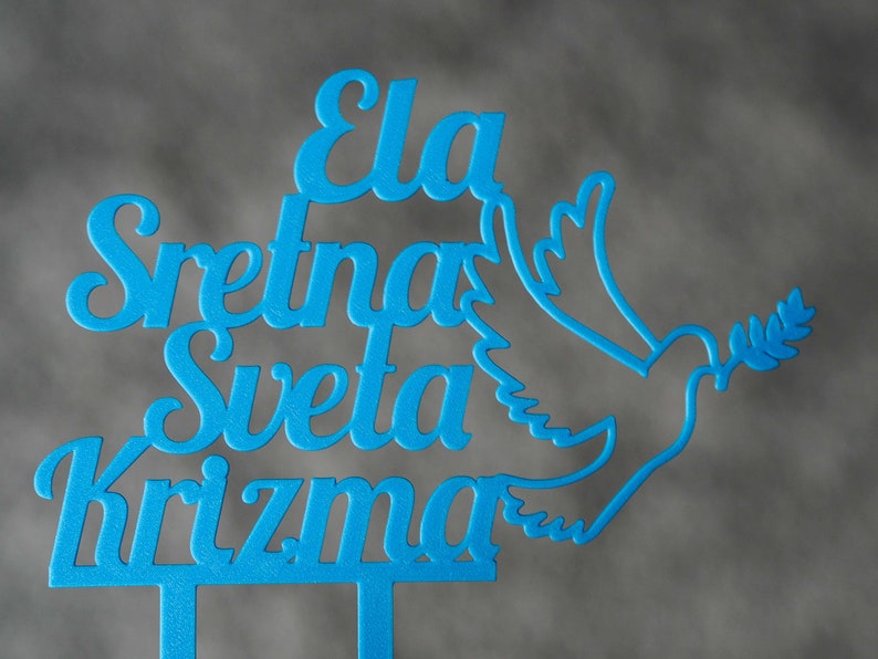 May include: A bright blue cake topper featuring the text "Ela Sretna Sveta Krizma" in a cursive font, accompanied by a stylized outline of a dove holding an olive branch. The design is cut from a single piece of material with two bottom prongs.