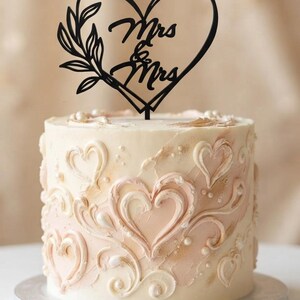May include: A black cake topper featuring a heart outline, a leafy branch, and the text Mrs & Mrs, positioned atop a cylindrical cake decorated with textured cream and pale pink frosting swirls, heart patterns, and small white pearl accents.