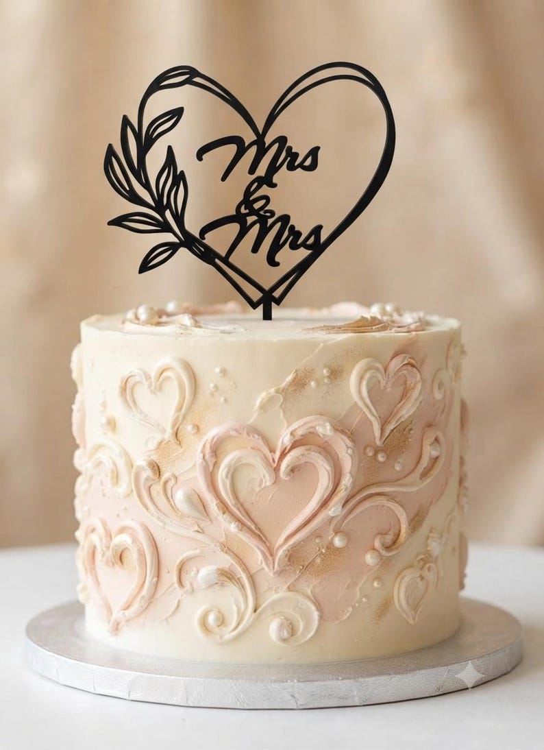 May include: A black cake topper featuring a heart outline, a leafy branch, and the text Mrs & Mrs, positioned atop a cylindrical cake decorated with textured cream and pale pink frosting swirls, heart patterns, and small white pearl accents.