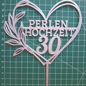 Może przedstawiać: Srebrny topper na tort w kształcie serca z gałązką z liśćmi po lewej stronie. Tekst PERLEN HOCHZEIT oraz liczba 30 znajdują się wewnątrz serca. Pojedynczy szpikulec wystaje od dołu, umożliwiając umieszczenie go w cieście.