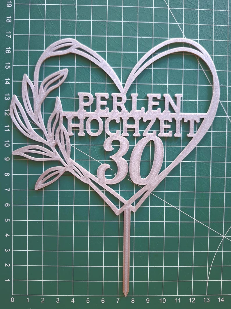 Może przedstawiać: Srebrny topper na tort w kształcie serca z gałązką z liśćmi po lewej stronie. Tekst PERLEN HOCHZEIT oraz liczba 30 znajdują się wewnątrz serca. Pojedynczy szpikulec wystaje od dołu, umożliwiając umieszczenie go w cieście.