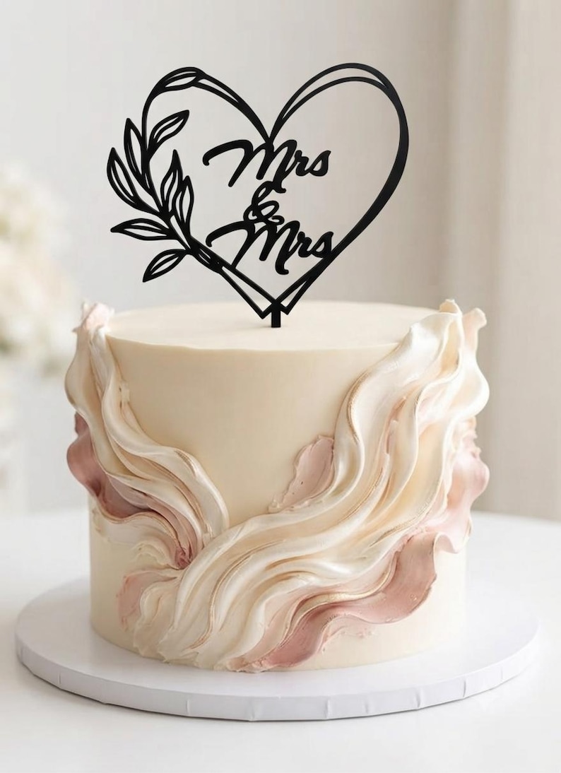 May include: A round cake with smooth cream-colored frosting and decorative vertical ruffles in shades of white and dusty rose. A black cake topper shaped like a heart with a leafy branch on the left side features the text Mrs & Mrs in script.