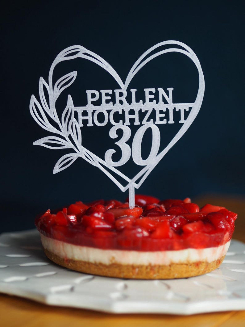 Może przedstawiać: Biały topper na tort w kształcie serca z gałązką liści po lewej stronie. Napis głosi PERLEN HOCHZEIT 30. Topper jest wbity w okrągły sernik truskawkowy ze złotym spodem i białą warstwą kremu.
