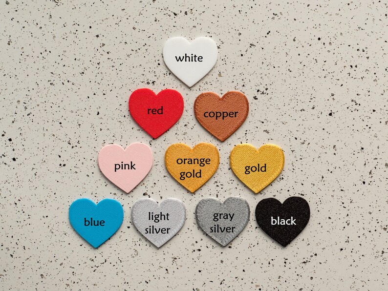 May include: A collection of ten heart-shaped cutouts arranged in a pyramid. The hearts are labeled with the colors white, red, copper, pink, orange gold, gold, blue, light silver, gray silver, and black, each featuring a distinct textured finish.