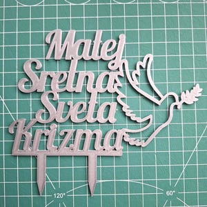 May include: A silver-colored cake topper featuring the text "Matej Sretna Sveta Krizma" in a cursive font, accompanied by a dove graphic holding an olive branch. The topper includes two pointed stakes at the bottom for insertion into a cake.