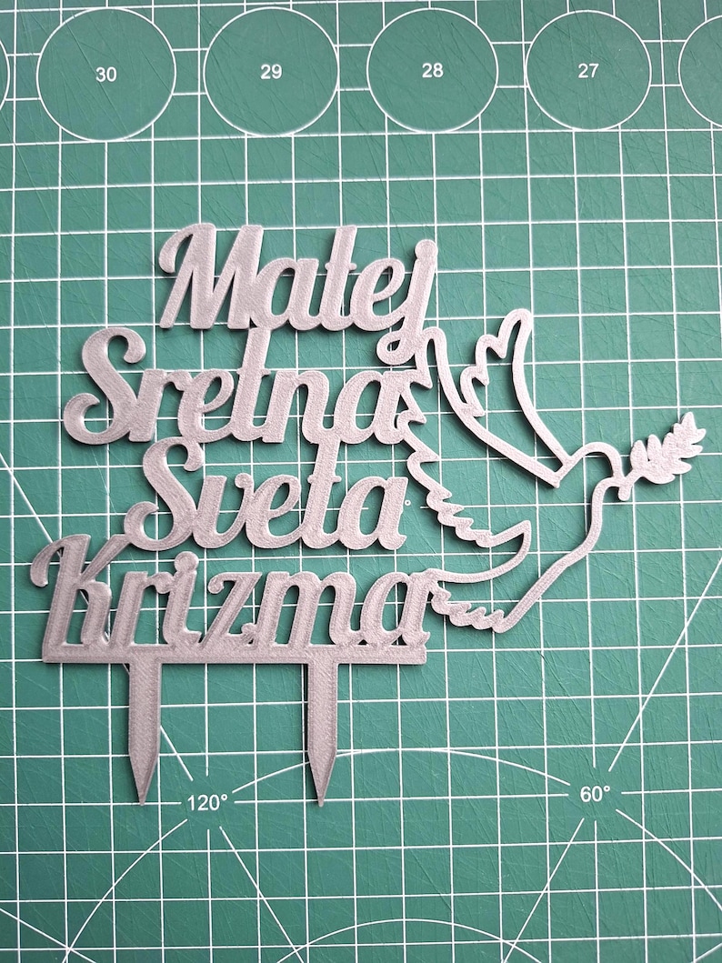 May include: A silver-colored cake topper featuring the text "Matej Sretna Sveta Krizma" in a cursive font, accompanied by a dove graphic holding an olive branch. The topper includes two pointed stakes at the bottom for insertion into a cake.