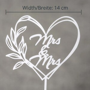 May include: A white cake topper in the shape of a heart with a leafy branch on the left side and the text Mrs & Mrs in the center. A measurement line at the top indicates a width of 14 cm. The topper has a single vertical stake at the bottom.