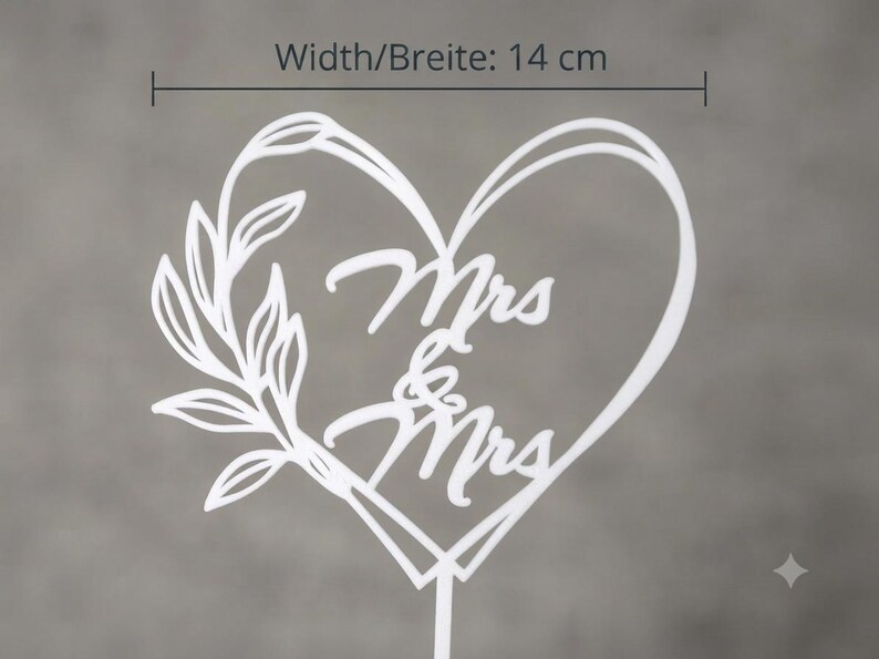 May include: A white cake topper in the shape of a heart with a leafy branch on the left side and the text Mrs & Mrs in the center. A measurement line at the top indicates a width of 14 cm. The topper has a single vertical stake at the bottom.