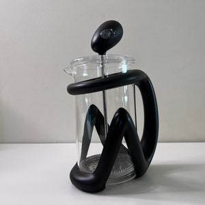Op de afbeelding: Een glazen cafetière met een zwarte handgreep en deksel. De cafetière heeft een cilindrische glazen kan en een zwart plunjermechanisme. De handgreep heeft een uniek, gebogen ontwerp.
