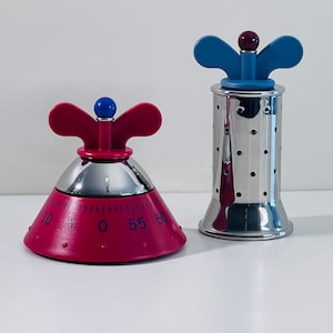 Op de afbeelding: Twee keukentimers. Eén is een magenta kegelvorm met een chromen basis en een blauwe knop. De andere is een lange, cilindrische timer van roestvrij staal met een blauwe en paarse knop. Beide hebben een vlindervormige handgreep.