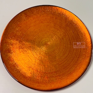 Puede incluir: Un plato de cena grande, redondo y de color naranja con un patrón de círculos concéntricos. El plato tiene un brillo metálico sutil y un borde ligeramente elevado. El plato está hecho de cerámica y tiene una etiqueta de marca que dice "IVV" en la esquina inferior derecha.