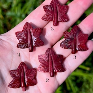 May include: Five dark red, carved stone maple leaf pendants, each featuring detailed vein textures and a central stem. The pieces are numbered 1 through 5, with each leaf displaying a unique, polished surface and deep crimson color variations.