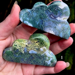May include: Two moss agate cloud-shaped carvings. The top carving is primarily blue and white with green accents. The bottom carving is green with white and blue accents. Both are held in a hand.