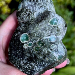 May include: A polished, irregular stone featuring a dark forest green and black mottled base with several smooth, circular inclusions of vibrant, translucent emerald green. The surface displays a natural, textured pattern with white mineral veins.