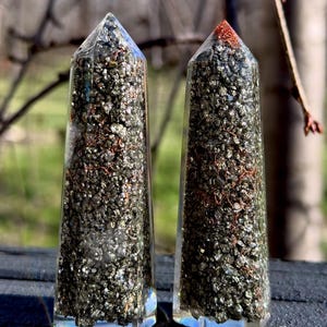 May include: Two clear, prism-shaped crystal obelisks filled with small, dark, metallic-looking stones and copper wire. The obelisks have a pointed top and a faceted base. One obelisk has a copper-colored top.