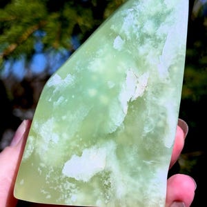May include: A polished, light green gemstone with white marbling. The stone has a smooth, rounded shape with a flat base and a pointed top. The gemstone is held in a hand, with a blurred background of green foliage.