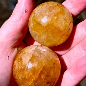 May include: Two golden-yellow crystal spheres, each with unique internal patterns, are held in a hand. The spheres have a smooth, polished surface and vary in color from light to dark amber. The spheres are numbered 1 and 2.