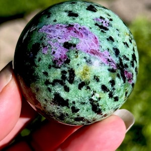 May include: A polished spherical stone featuring a vibrant green base color with prominent black mineral inclusions and distinct patches of deep magenta-pink crystalline formations distributed across the smooth, rounded surface of the orb.