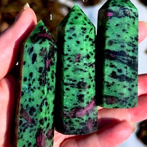 May include: Three green zoisite crystal towers with black speckles and red ruby inclusions. Each tower has a pointed top and a rectangular base. The crystals are held in a hand, with the background out of focus.