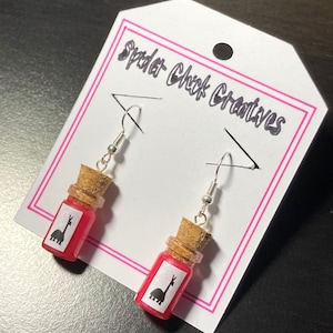 May include: A pair of earrings featuring small glass bottles filled with pink liquid and a black silhouette of a llama. The bottles have cork stoppers and are attached to silver earring hooks. The earrings are displayed on a white card with the text "Spider Chick Creatives".