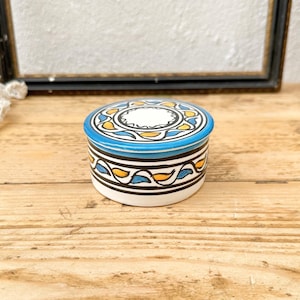 May include: A small, round ceramic trinket box with a lid. The box features a white base, a band of black, yellow, and blue geometric patterns, and a blue lid with a white and yellow floral design. The box sits on a wooden surface.