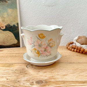 May include: A cream ceramic plant pot with a matching saucer, decorated with a floral pattern in pink, yellow, and green. The pot has a scalloped edge with a pale blue trim. The set sits on a wooden surface.