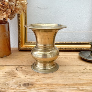May include: A vintage brass vase with a flared top and a rounded base. The vase is displayed on a rustic wooden surface, with a gold-framed picture and dried flowers in the background. The vase is approximately 15 cm tall.