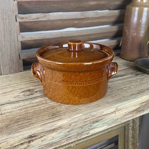 May include: A brown ceramic pot with a lid and two handles. The pot is sitting on a wooden shelf.