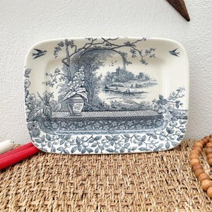 May include: Rectangular ceramic tray with a blue and white pastoral scene. The tray features a detailed landscape with trees, buildings, and figures. The border has a floral design. Two birds are depicted on the tray.