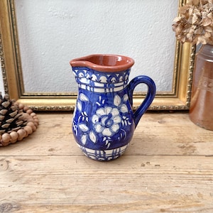 May include: A small ceramic pitcher featuring a deep blue glaze with hand-painted white floral motifs and leaf patterns. The vessel has a rounded body, a curved handle, and a wide pouring spout with a natural terracotta-colored rim and interior.
