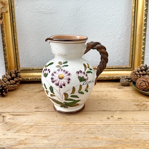May include: Cream-colored ceramic pitcher with a brown braided handle and rim. The pitcher is decorated with purple and white floral designs and green leaves. The word "cicoria" is written on the pitcher. The pitcher sits on a wooden surface.