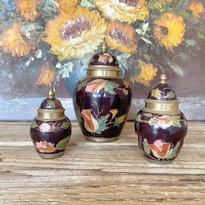 May include: Three decorative brass jars with lids. The jars are painted with a dark purple background and floral designs in shades of pink, green, and yellow. The jars are arranged in a row on a wooden surface.