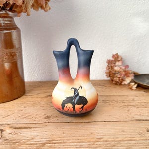 May include: A small, two-handled vase with a unique shape. The top is dark blue, transitioning to orange and white with a painted scene of a figure on horseback. The vase is displayed on a wooden surface.